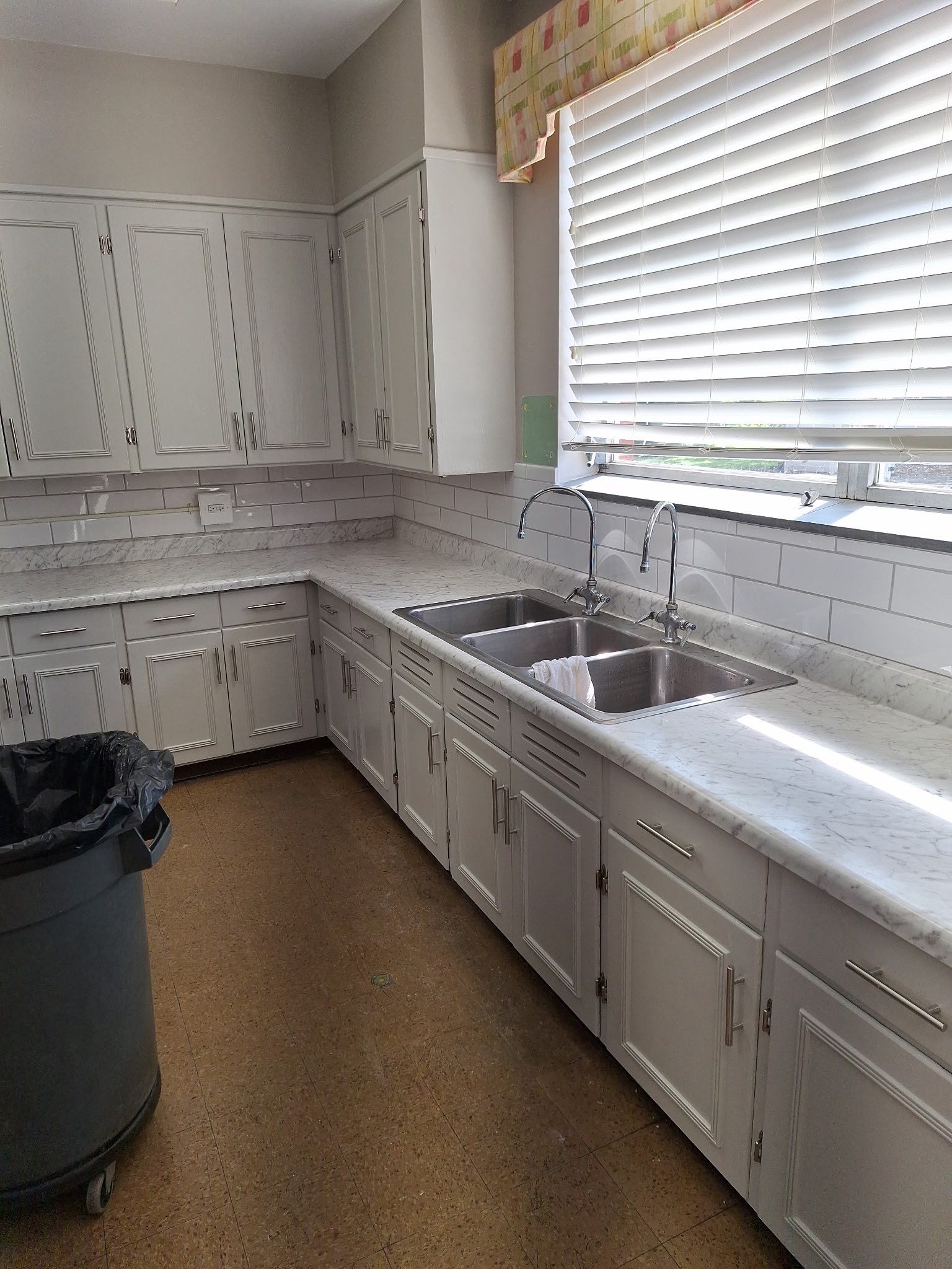 Kitchen with white cabinets, countertops, and backsplash, stainless steel sink, and window with blinds.
