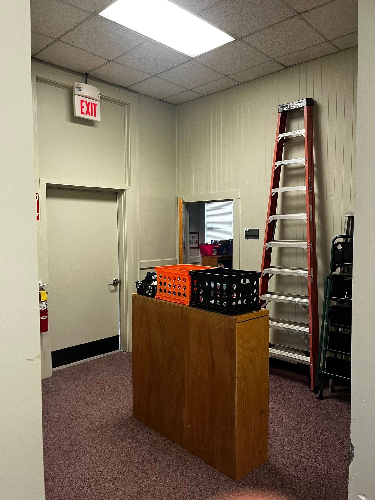 An interior shot of a storage room with a wooden table, crates, a ladder, and an exit sign.