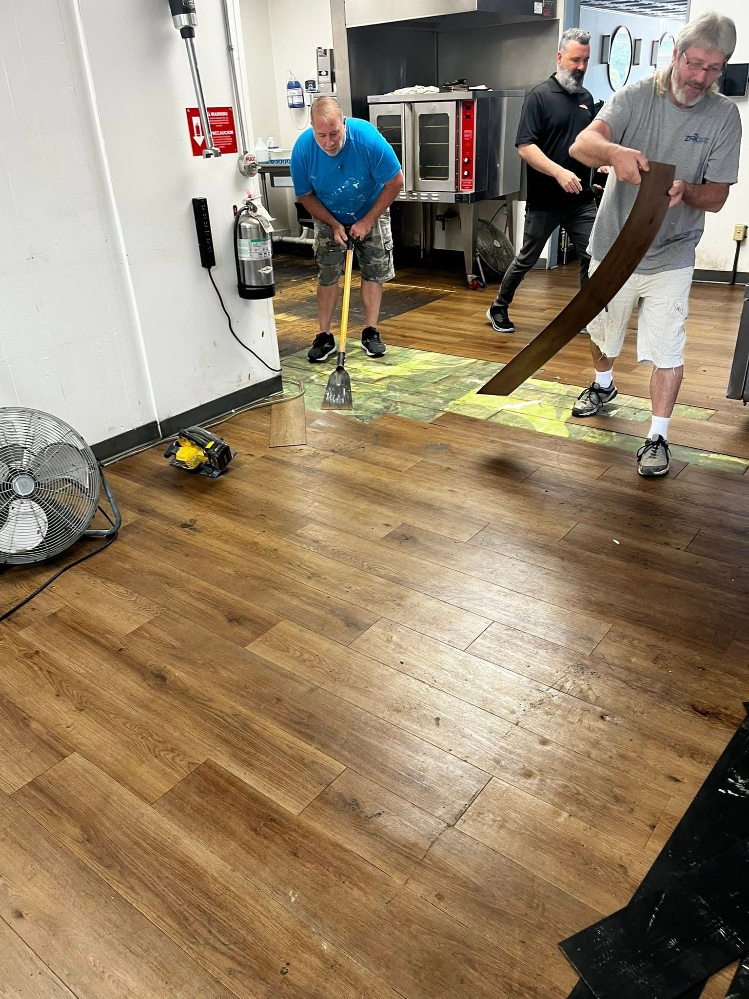 Men cleaning up spilled green substance in a kitchen. One swings a golf club, another uses a board.