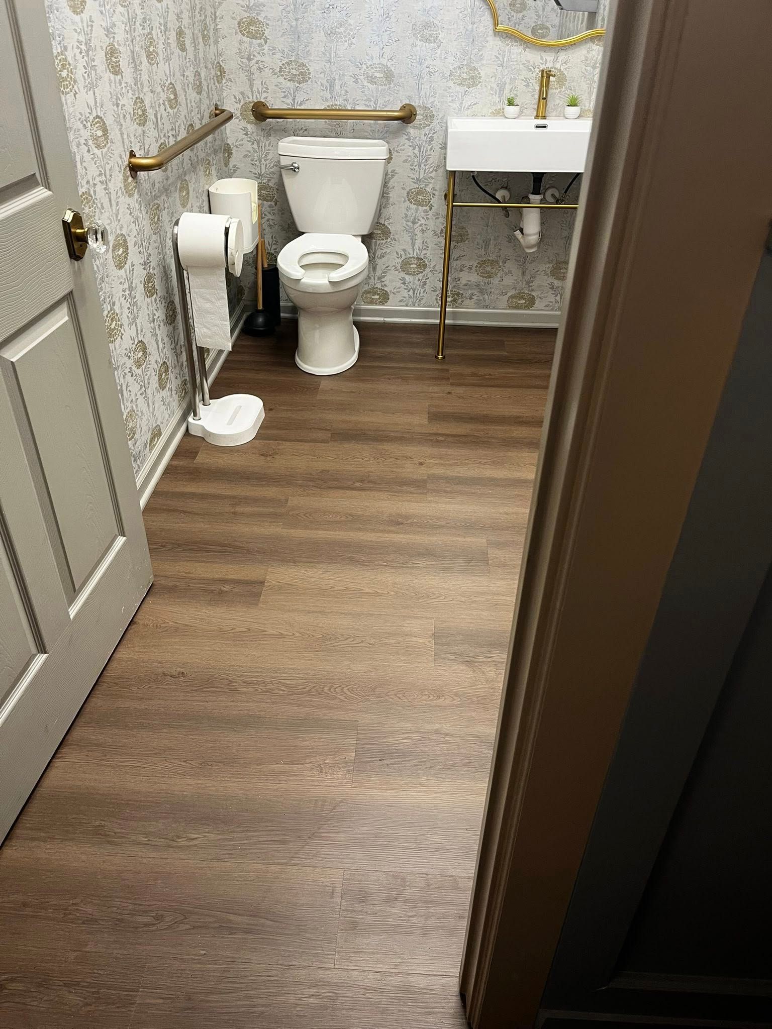 Small bathroom with wood-look flooring, toilet, sink, and decorative wallpaper; open doorway on the right.