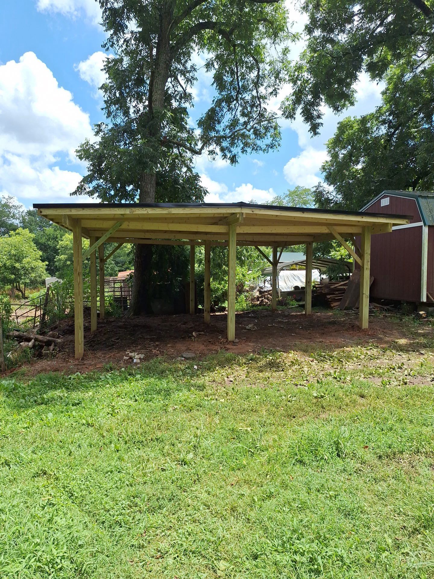 Wooden carport with black roof, in a grassy backyard, under a large tree.