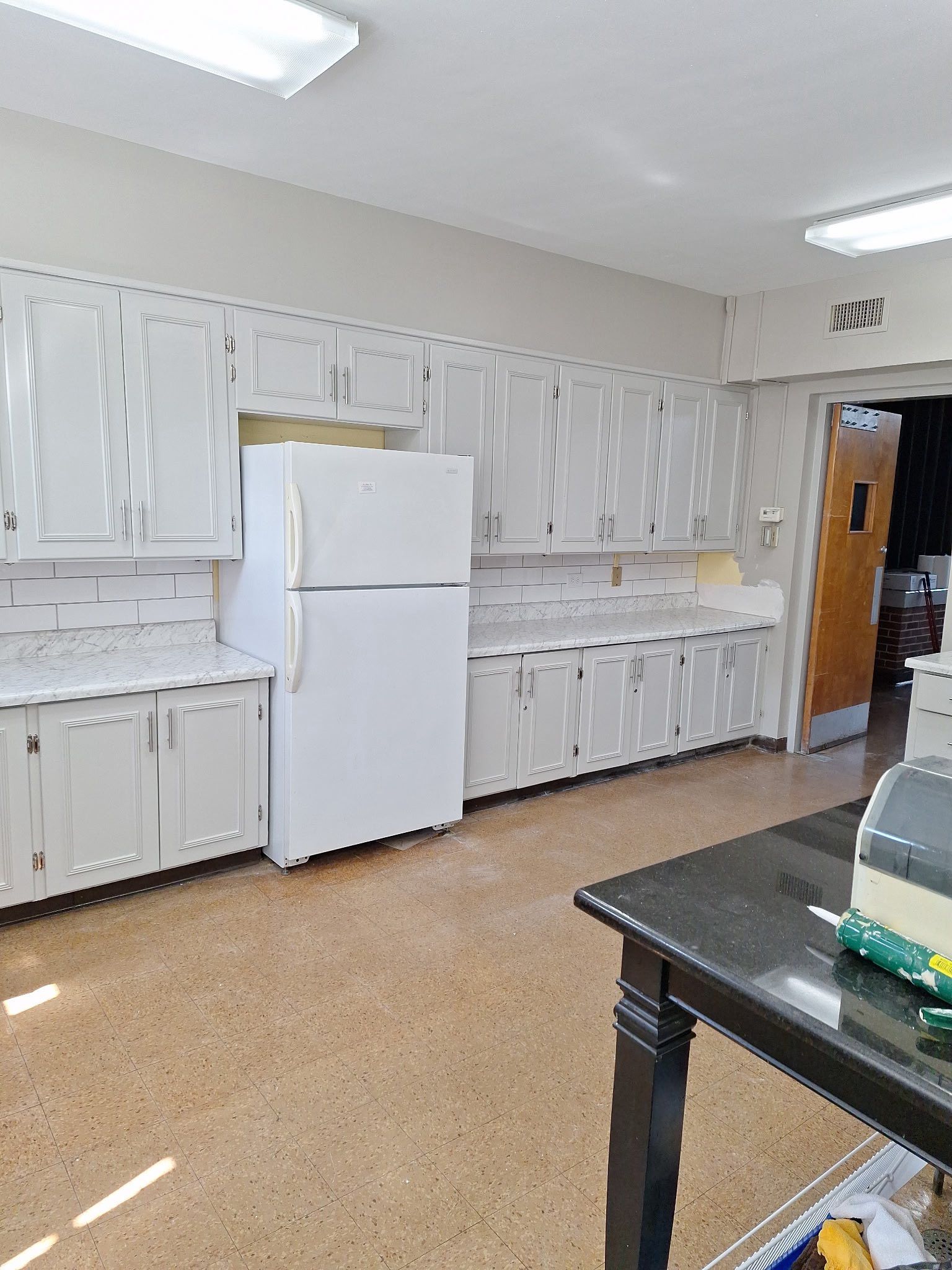 White kitchen with cabinets, countertops, and a refrigerator. A dark table is in the foreground.