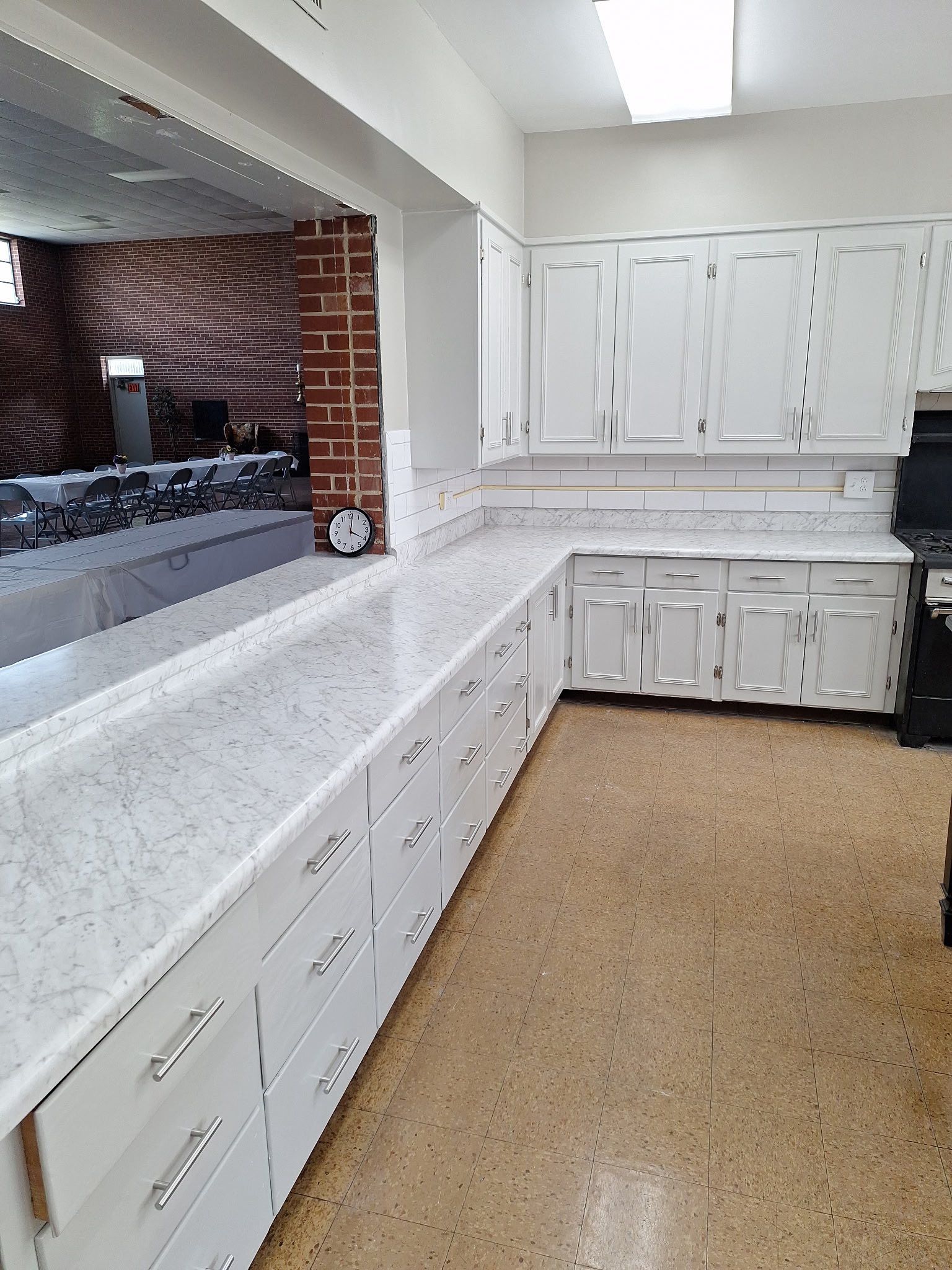 White kitchen cabinets and countertops in a room with a pass-through window and brick wall.