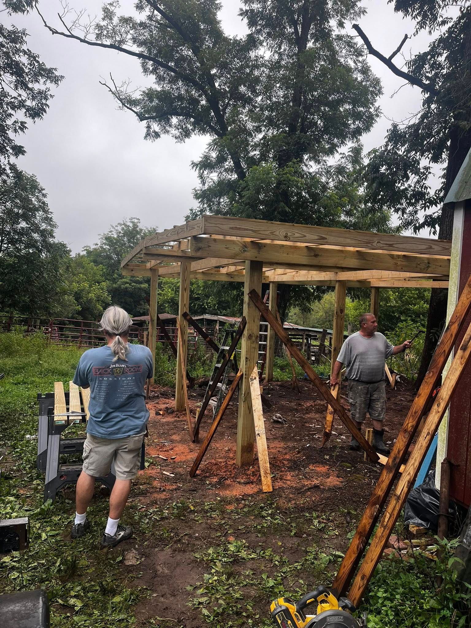 Two people building a wooden structure outdoors. One takes a photo, the other holds a beam. Overcast, muddy ground.
