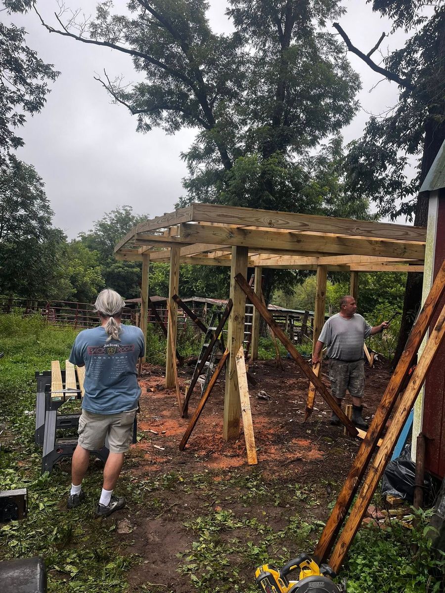 Two men building a wooden structure outdoors. One takes a photo, the other holds a beam. Overcast, muddy ground.