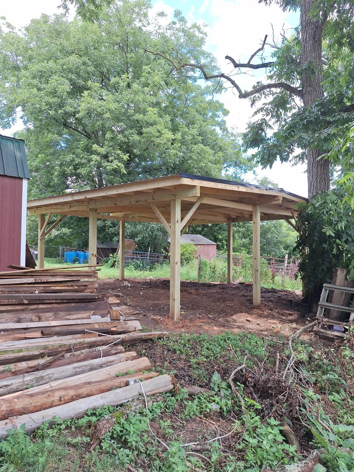 Wooden pavilion under construction in a rural setting, with lumber and trees nearby.