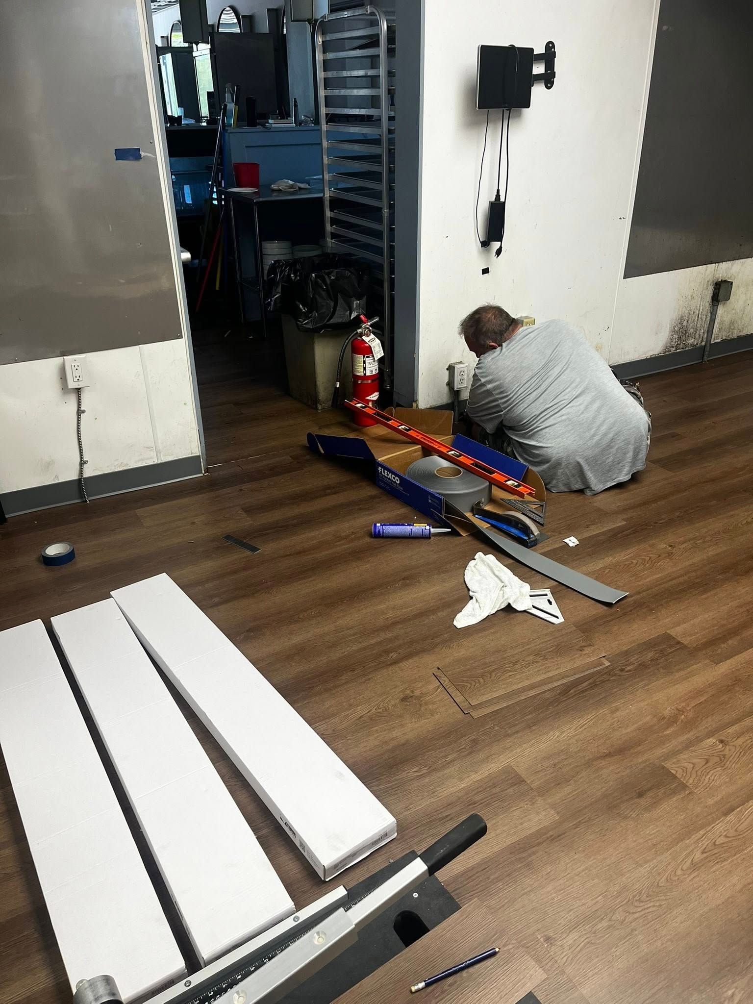 Person kneeling, working on floor near tools and materials in a commercial space with wood-look flooring.