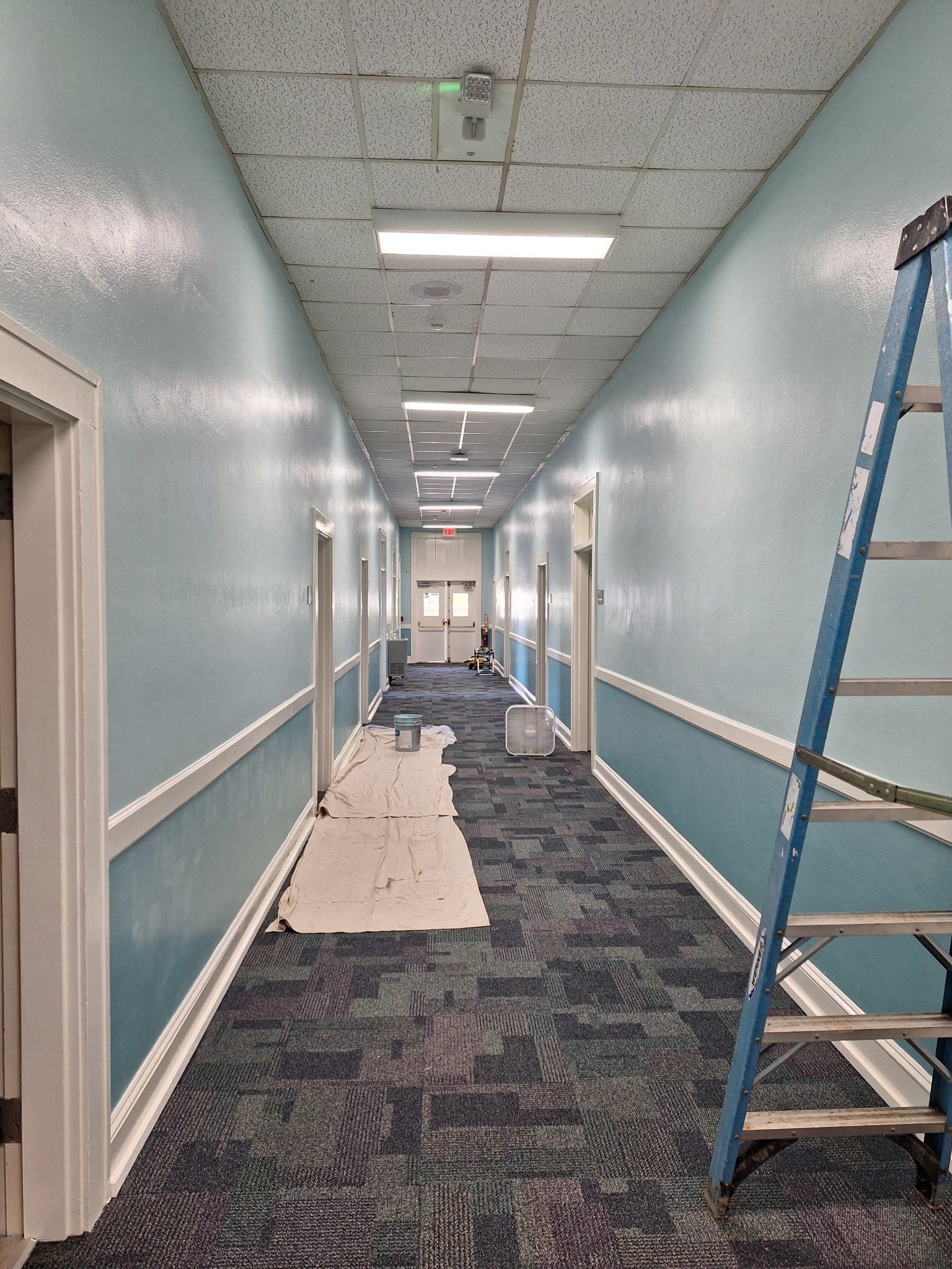 Long hallway painted light blue, white trim, with ladder and paint supplies.