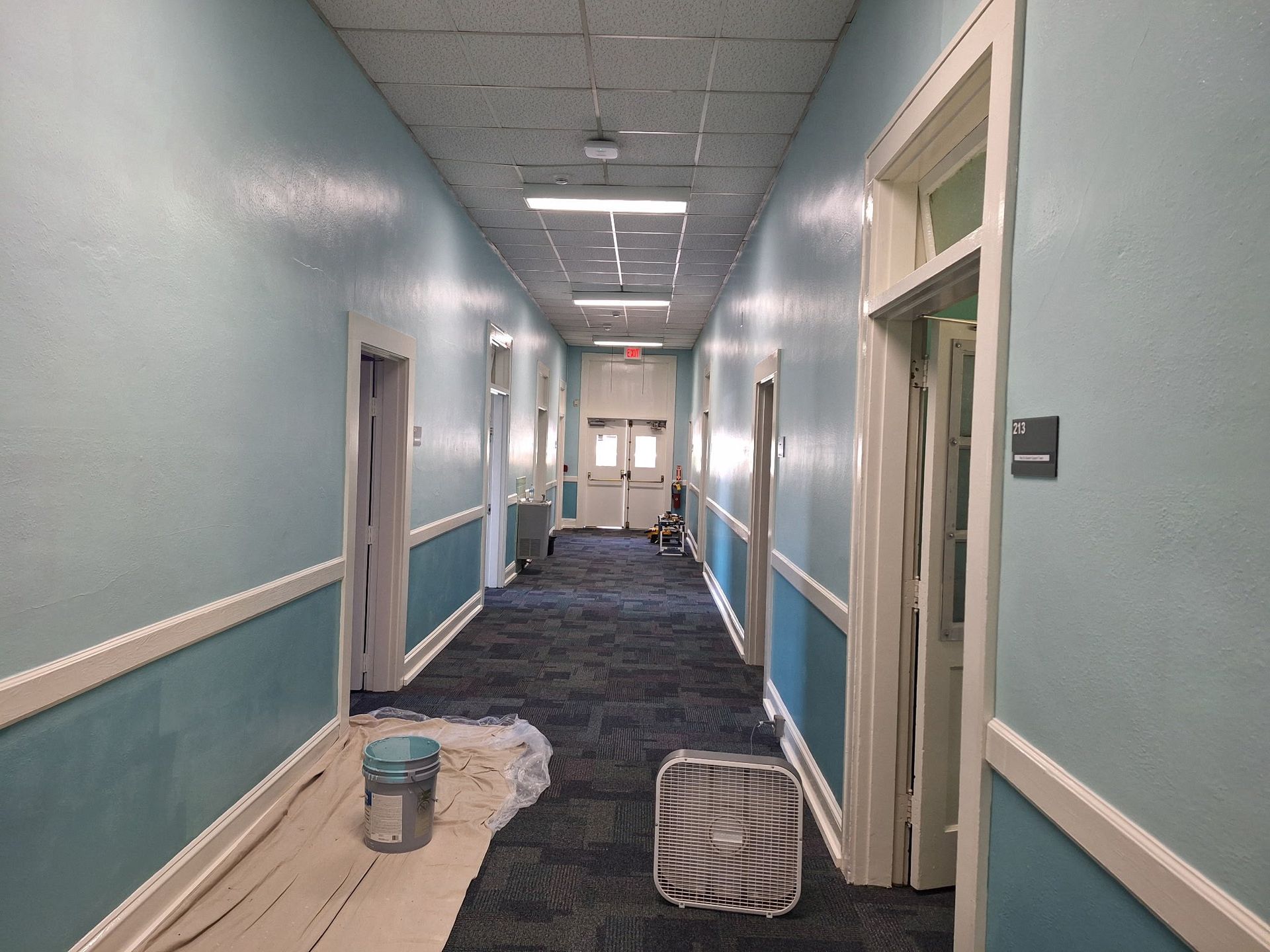 Long hallway with blue walls, white trim, and a patterned carpet. A fan and paint bucket are visible.