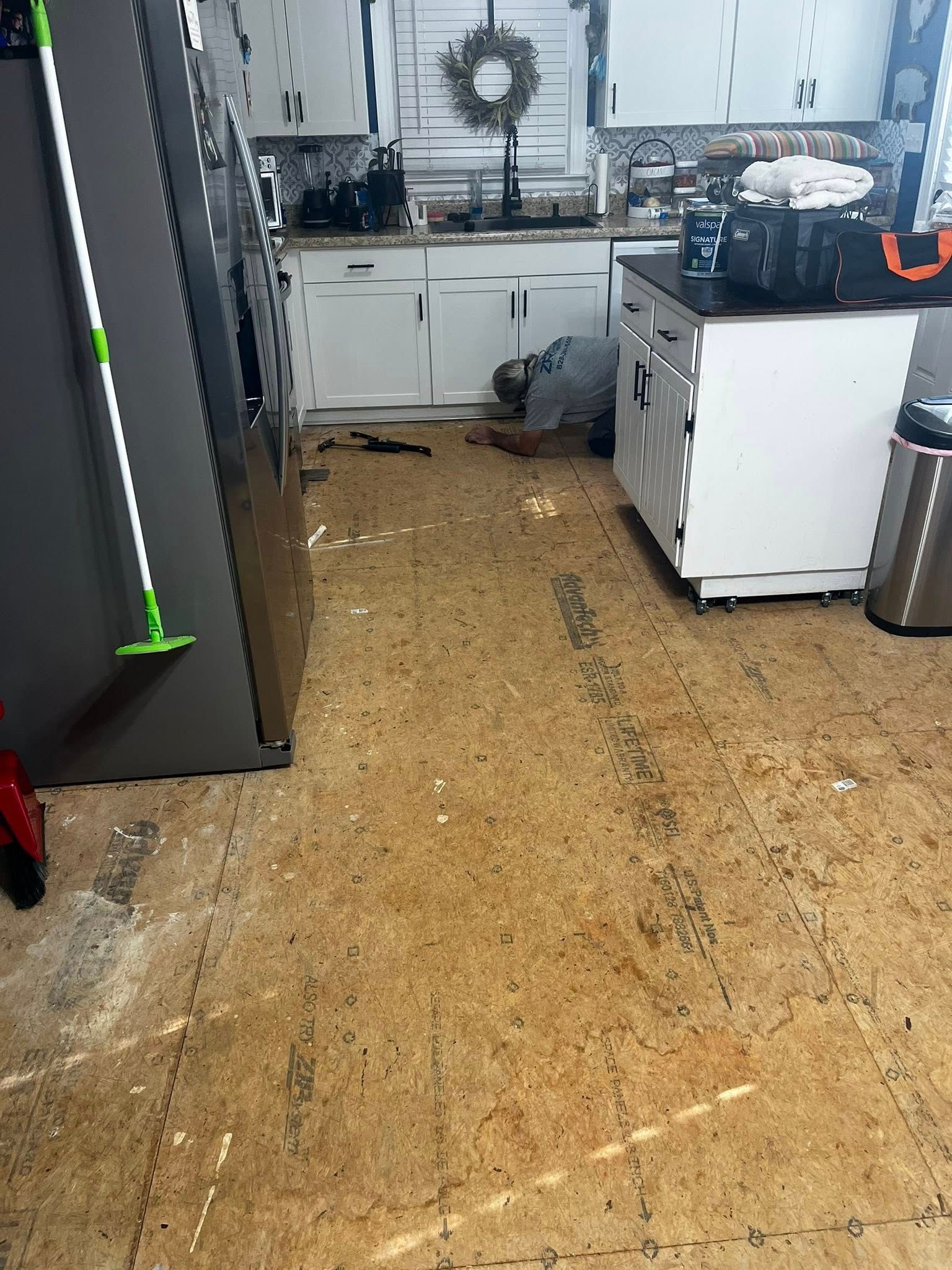Person working on floor of kitchen with white cabinets, beige flooring, and a stainless steel refrigerator.