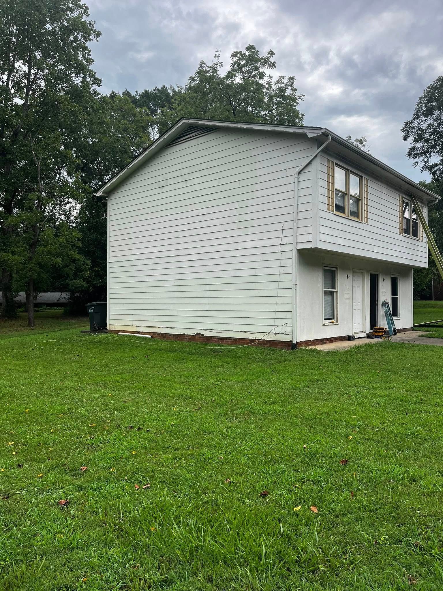 Two-story white house with damaged siding under an overcast sky, surrounded by green grass and trees.