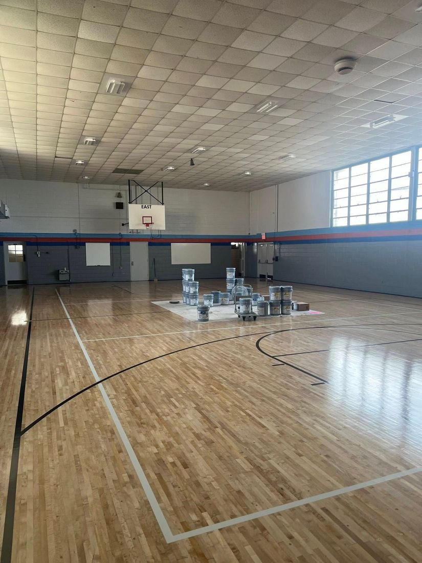 Gymnasium with wooden floor, basketball hoop, and gray walls, with a grid-patterned ceiling and daylight streaming through the windows.