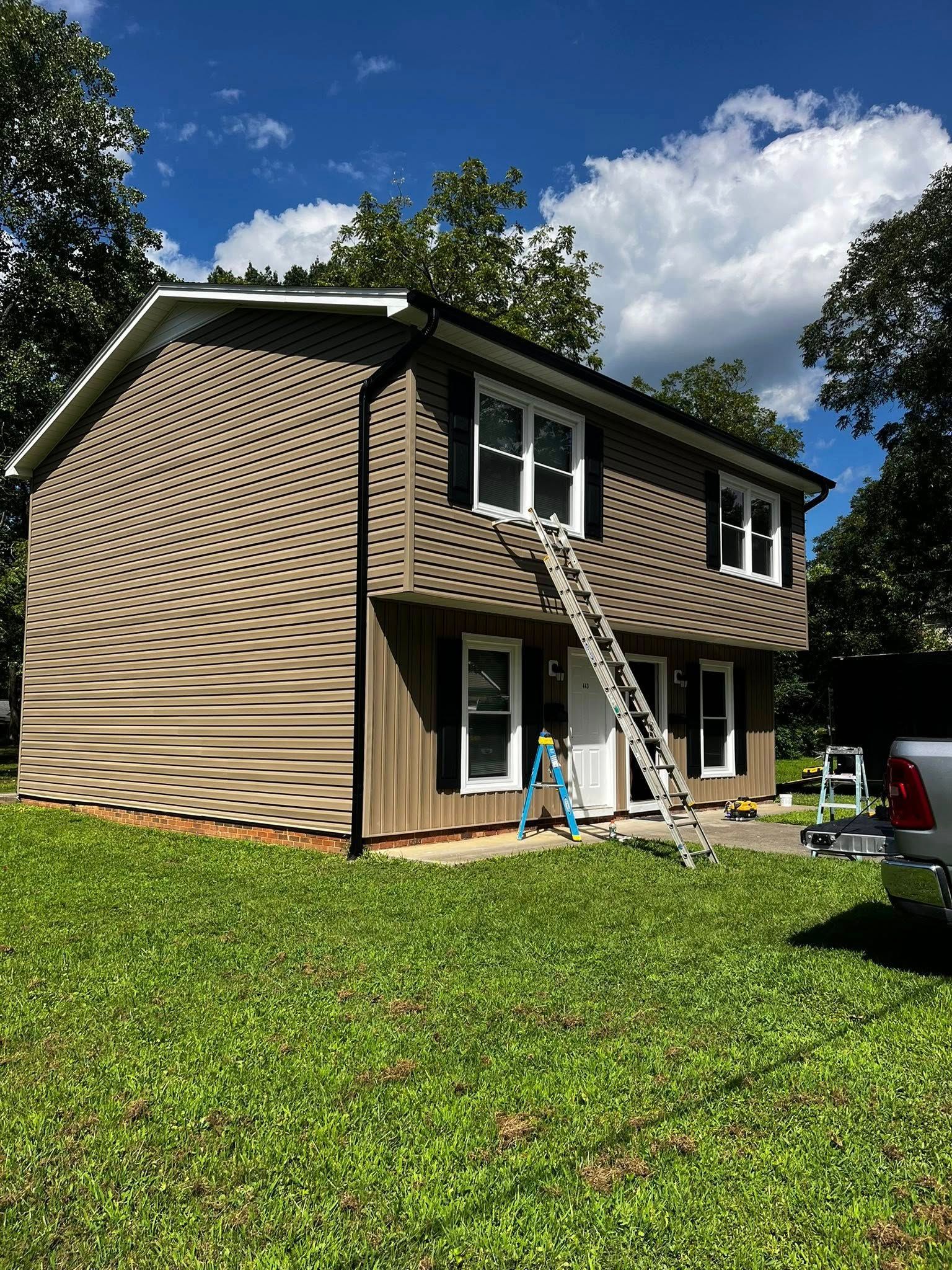 Two-story house with brown siding, white windows, and a ladder leaning against it on a grassy lawn.