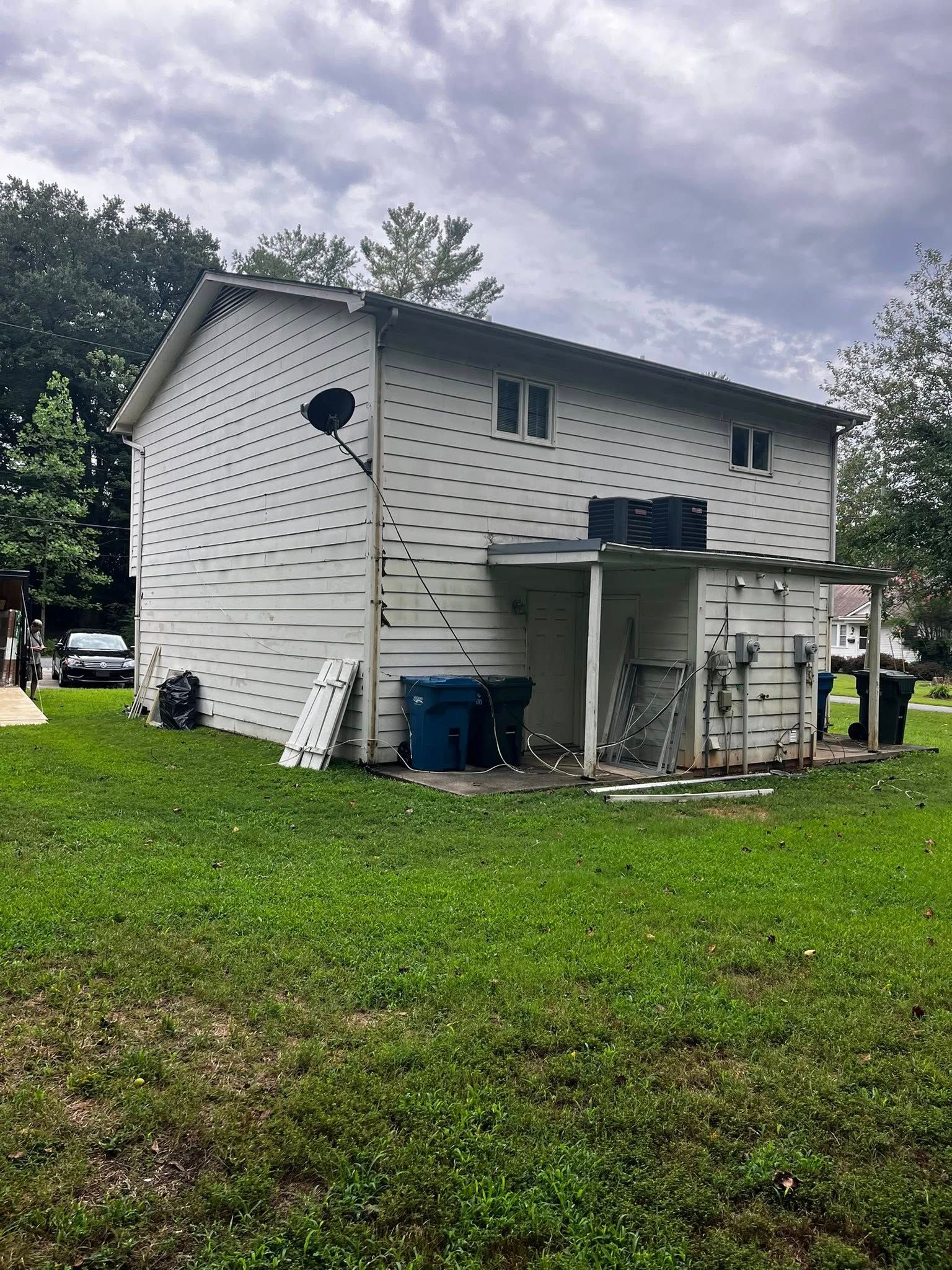 Back of a two-story white house with a covered area. Blue trash cans sit on the grassy lawn under a cloudy sky.