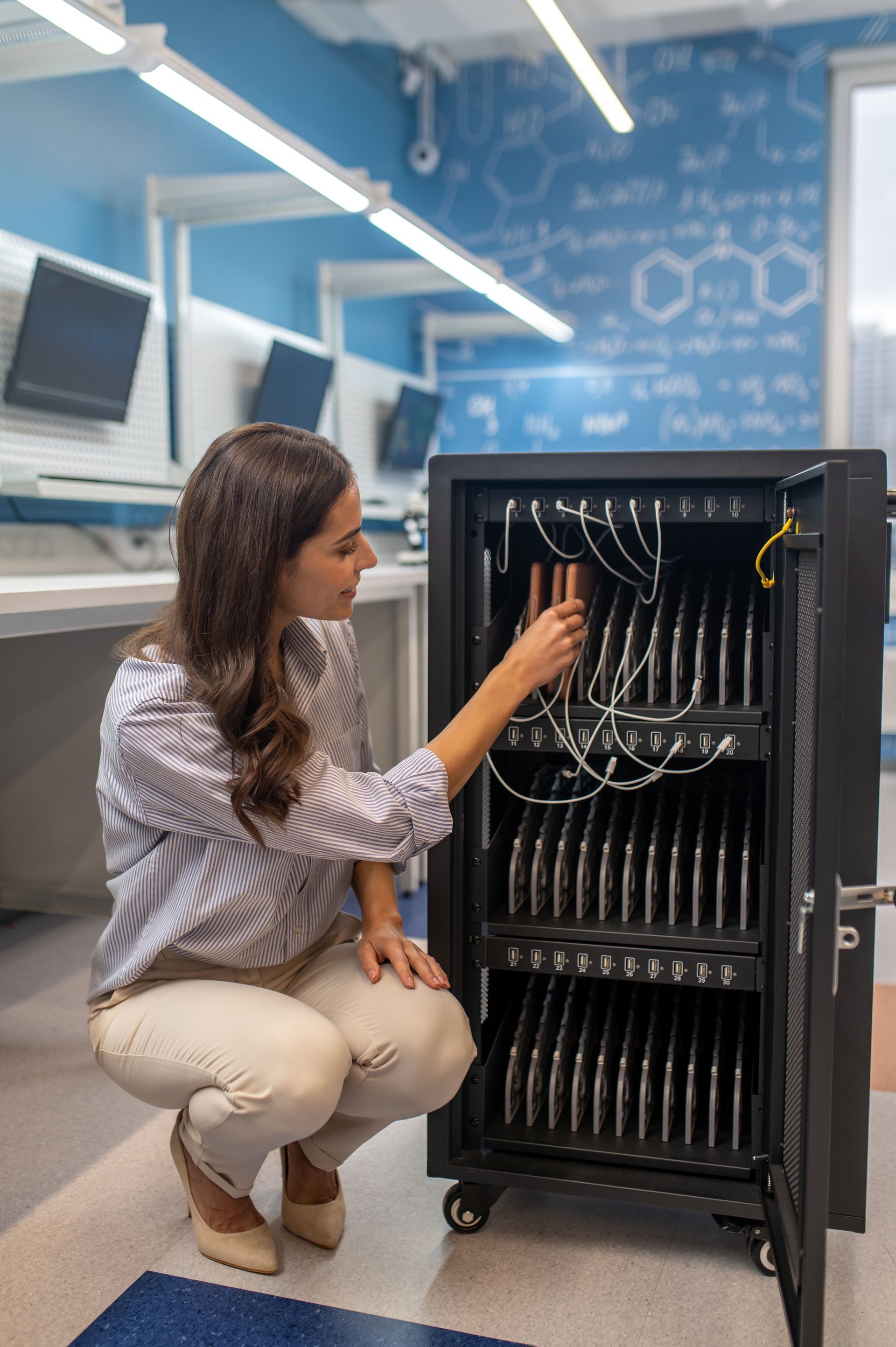 A woman is kneeling down in front of a cabinet filled with laptops.