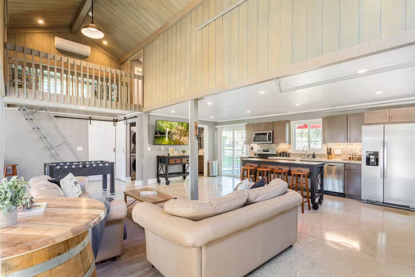 Open-plan living space with kitchen, lounge area, and a loft. Exposed beams, light walls, and a glossy floor.