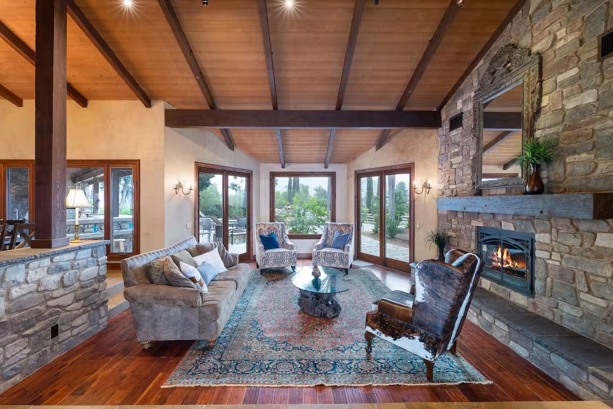 Living room with stone fireplace, wood ceiling, windows, and plush furniture.