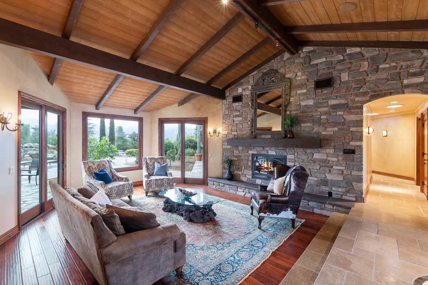 Living room with stone fireplace, wooden ceiling, and large windows overlooking a garden.