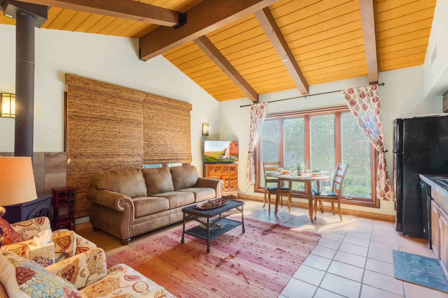 Cozy living room with brown sofa, rug, and dining table near a window. Wooden ceiling.