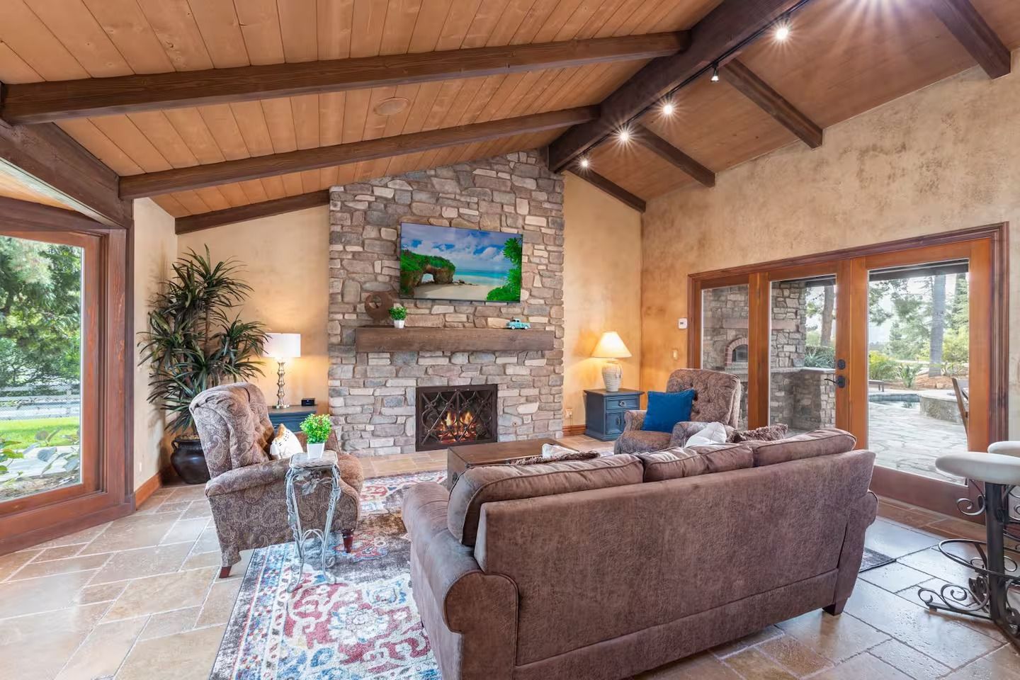 Living room with stone fireplace, brown furniture, wooden ceiling, and large windows.
