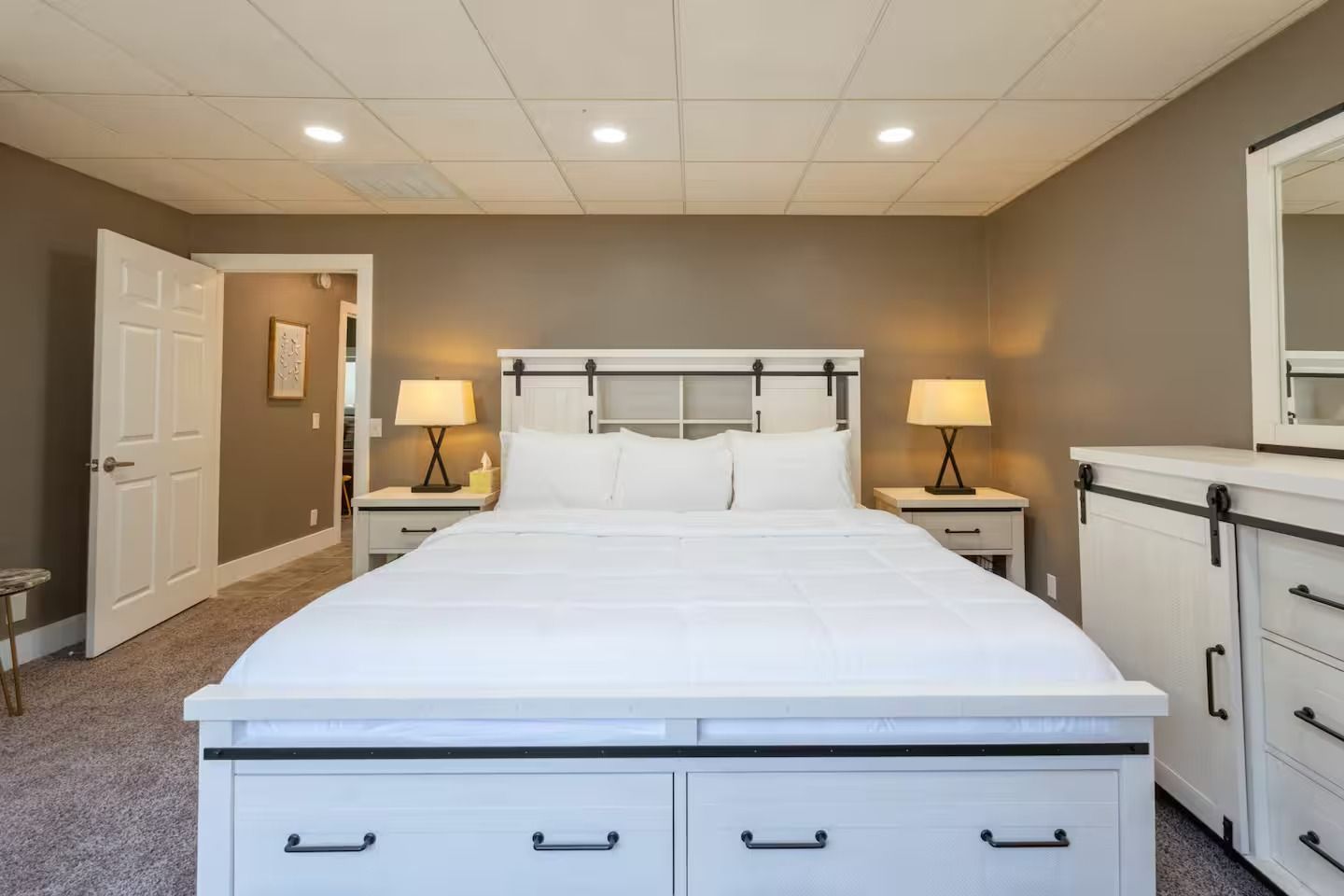 Bedroom with white bed, nightstands, dresser. Beige walls, carpet, door, lamps, and ceiling lights.
