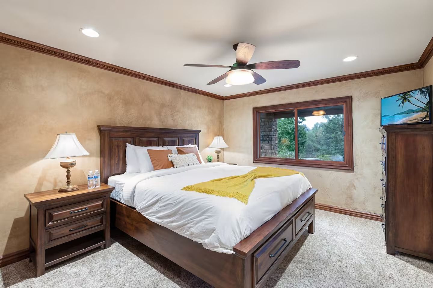 Bedroom with a wooden bed, nightstands, and dresser. Light-colored walls and carpet. Window with a view.