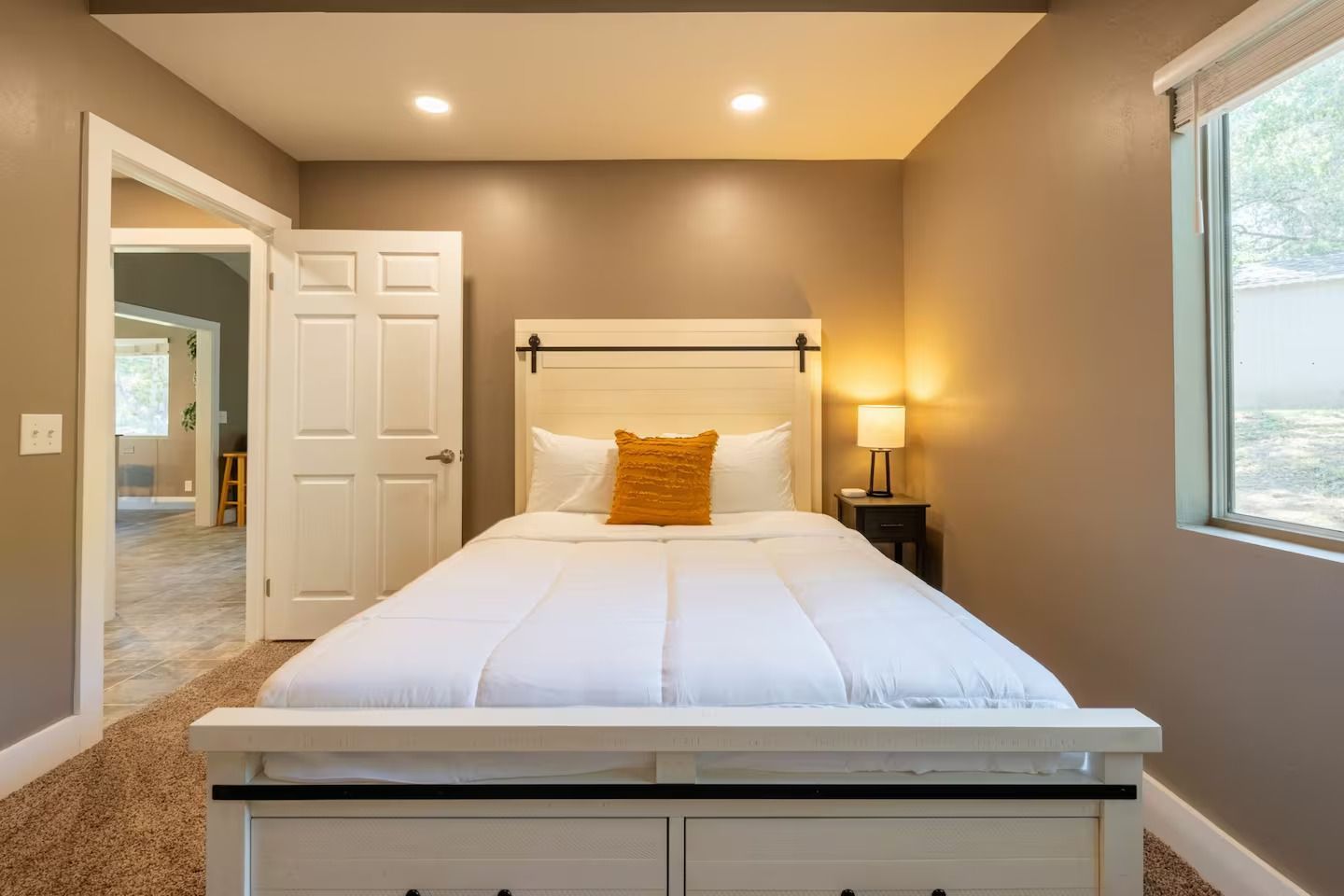 Bedroom with a white bed, beige walls, open door to hallway, and window with greenery view.