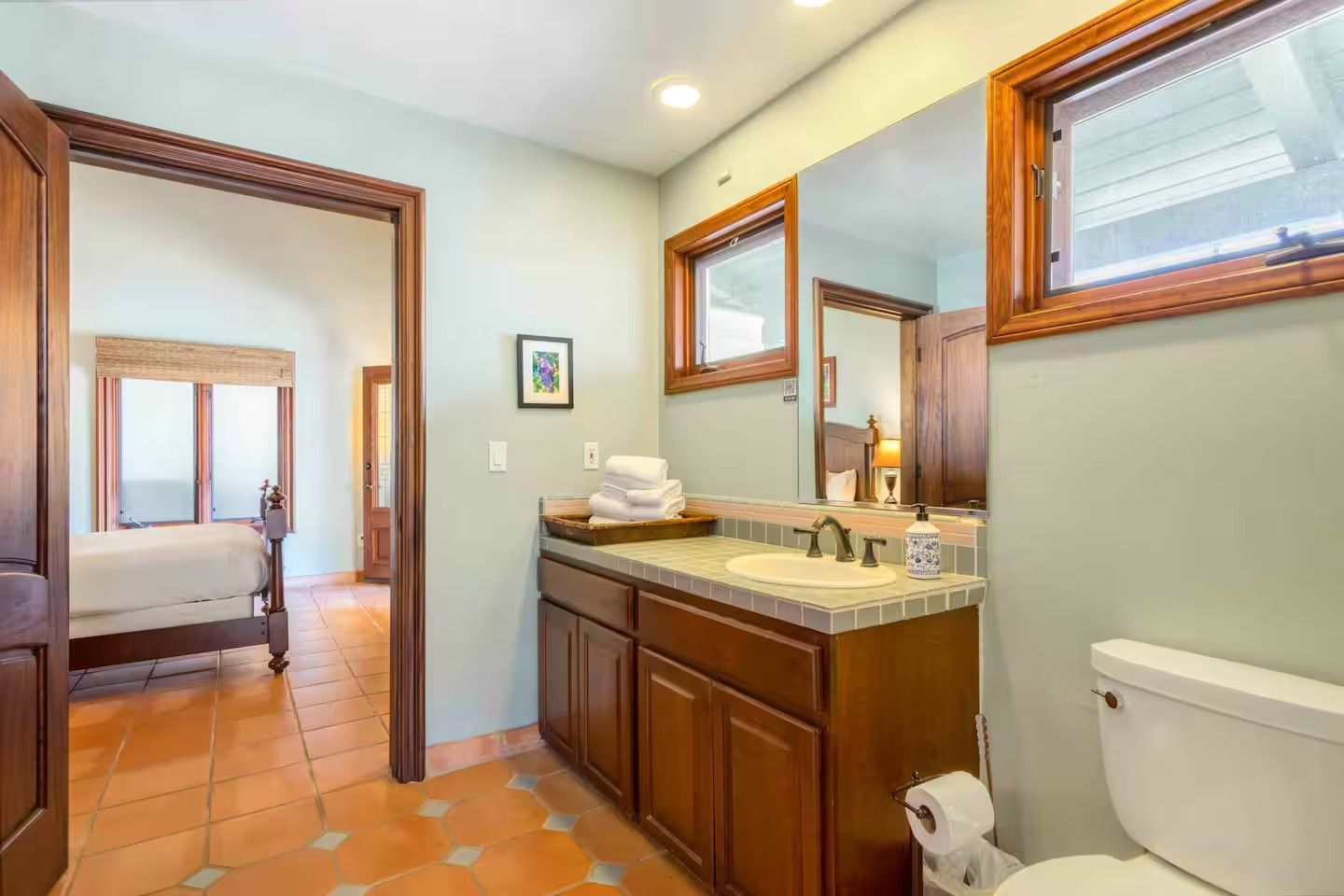 Bathroom with brown vanity, toilet, and doorway to a bedroom with bed.