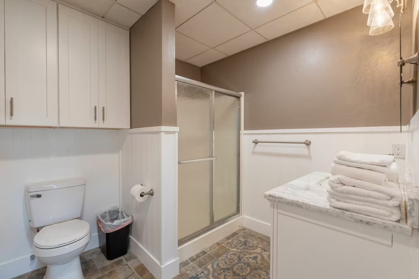 Bathroom with toilet, vanity, shower, and stacked towels; white and brown tones.