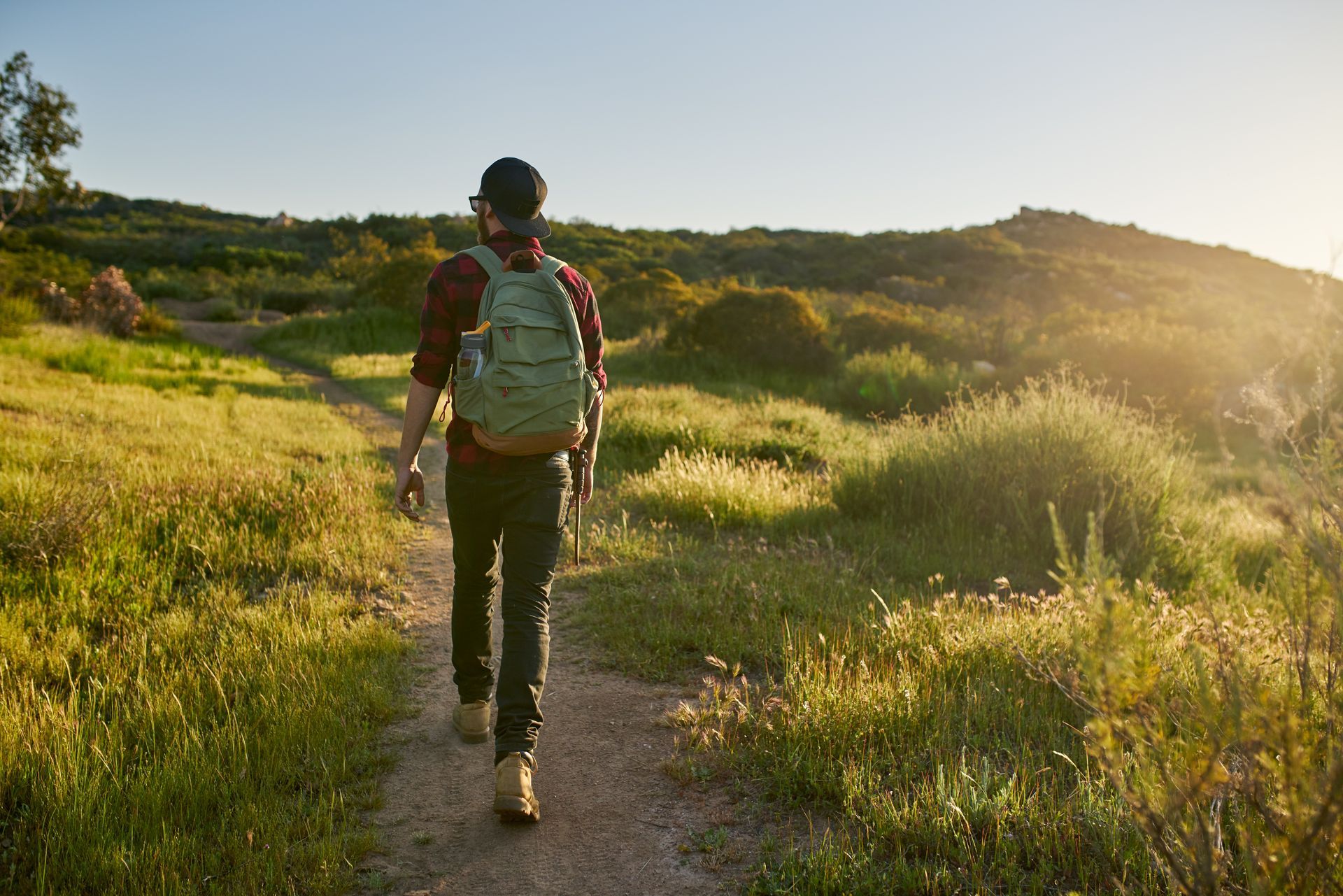Hiker walking a sunlit trail through green hills, wearing a backpack and holding a water bottle.