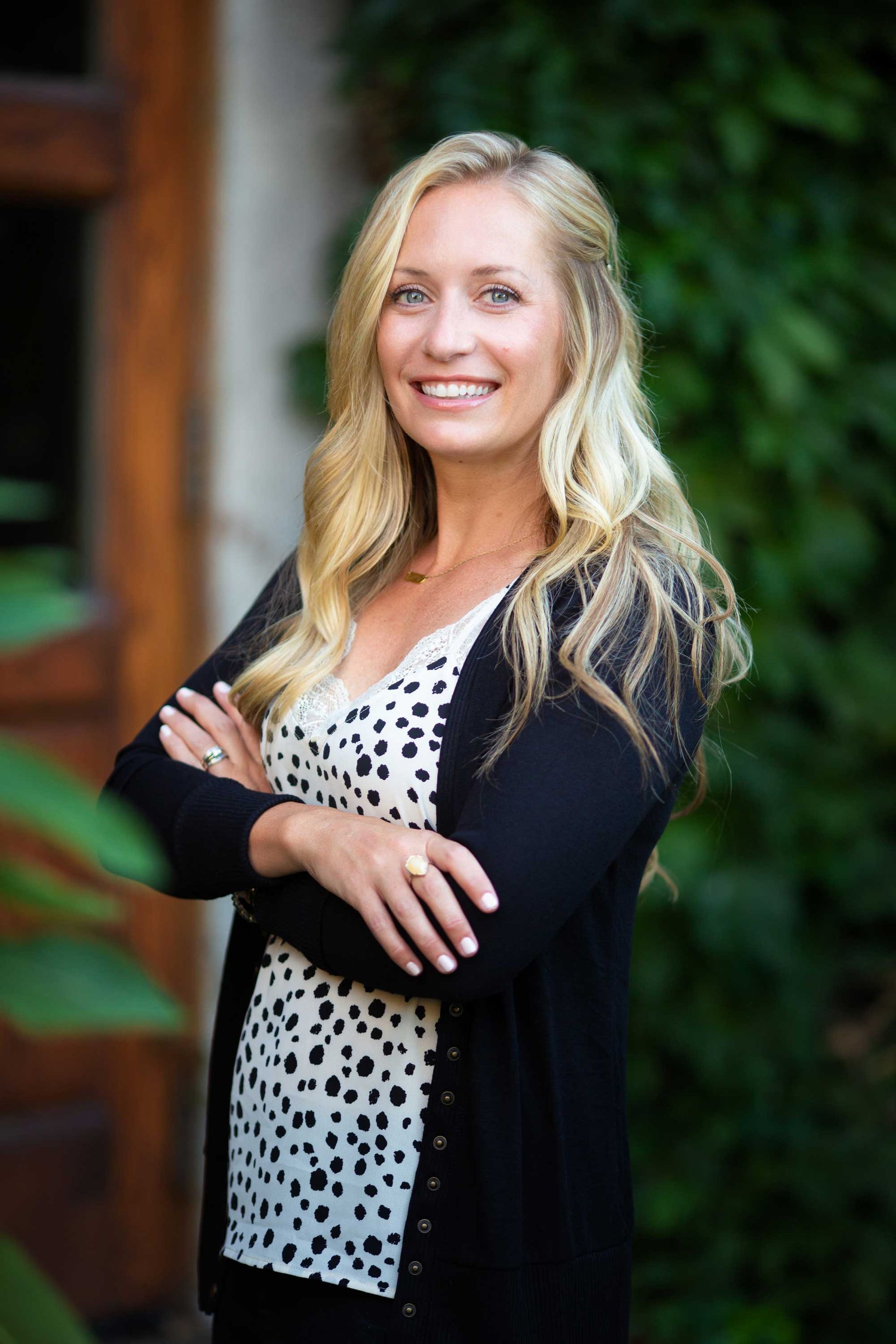 Woman with blonde hair, arms crossed, smiling. Wearing black sweater, spotted shirt, outdoors. Woman with blonde hair, arms crossed, smiling. Wearing black sweater, spotted shirt, outdoors.