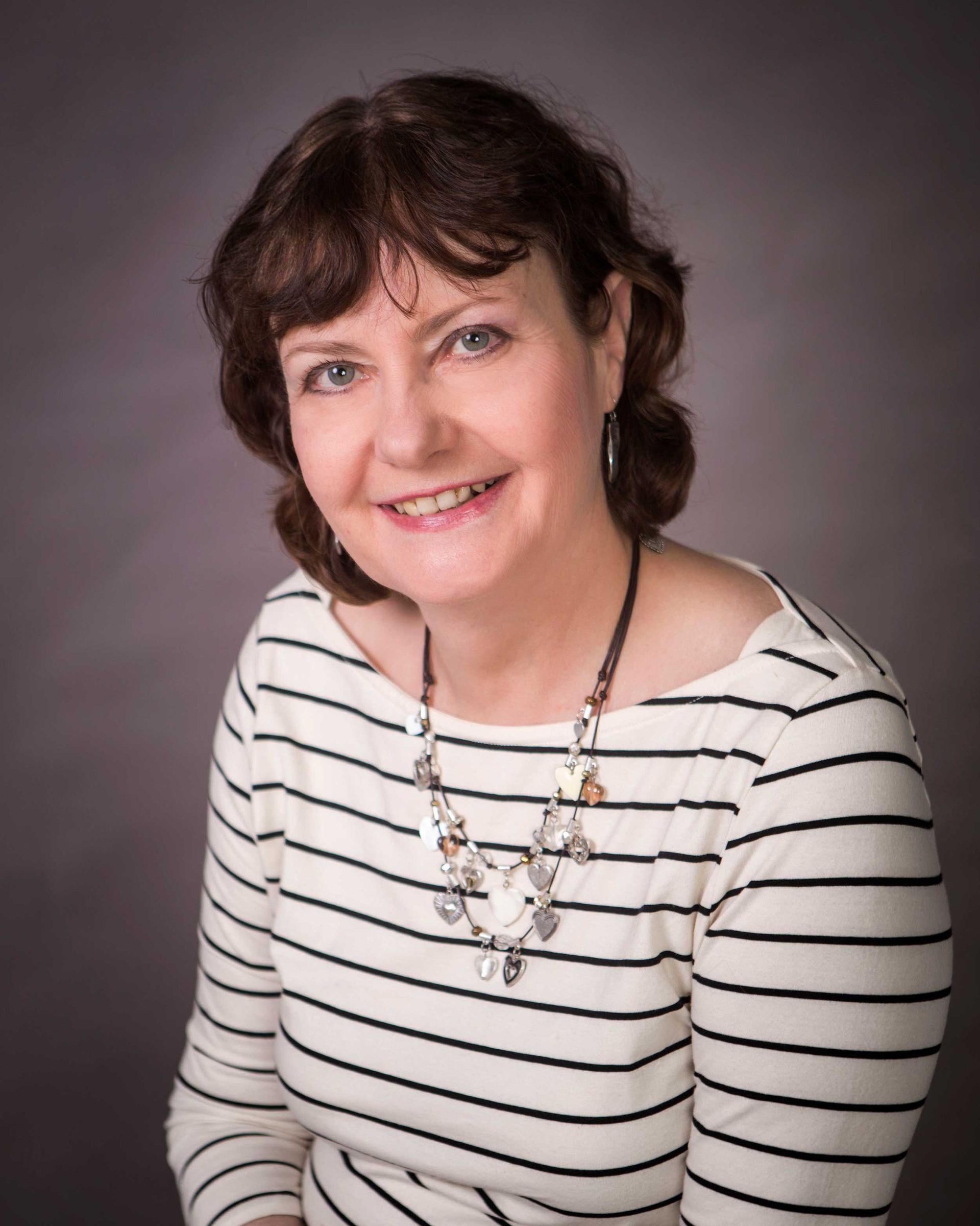 Woman with brown hair smiles, wearing a striped top and necklace, posing against a gray background. Woman with brown hair smiles, wearing a striped top and necklace, posing against a gray background.