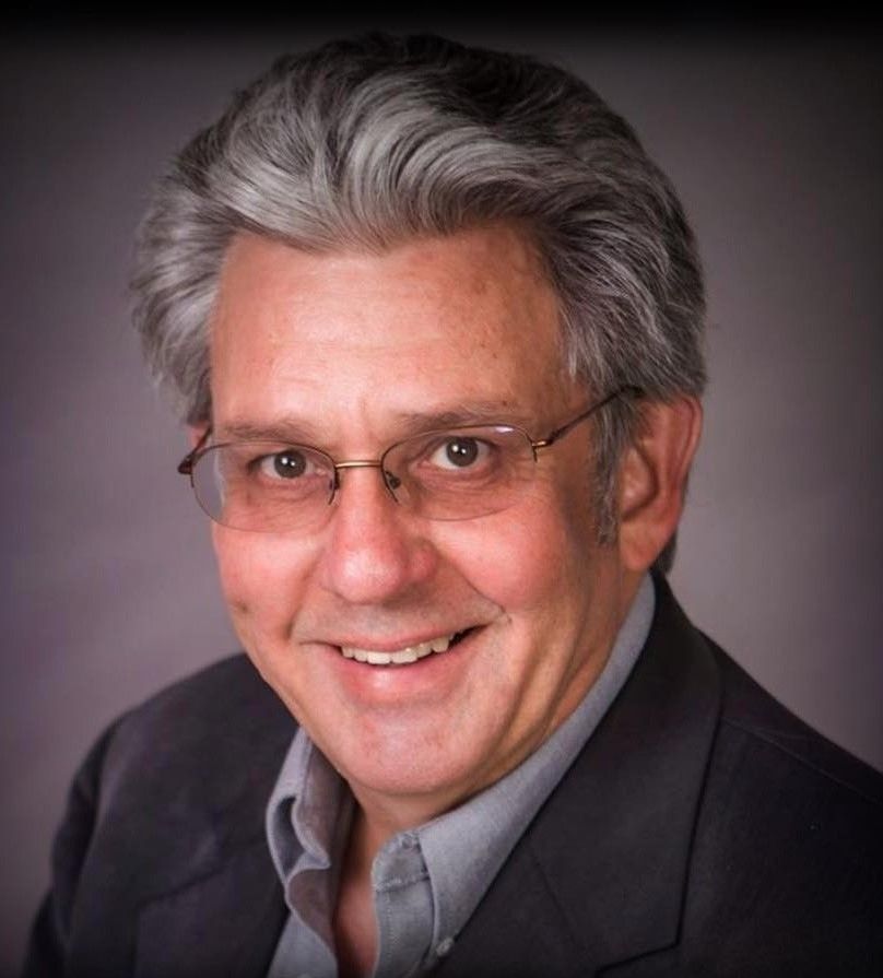Man with glasses smiling, wearing a gray blazer and light blue shirt. Gray hair. Man with glasses smiling, wearing a gray blazer and light blue shirt. Gray hair.