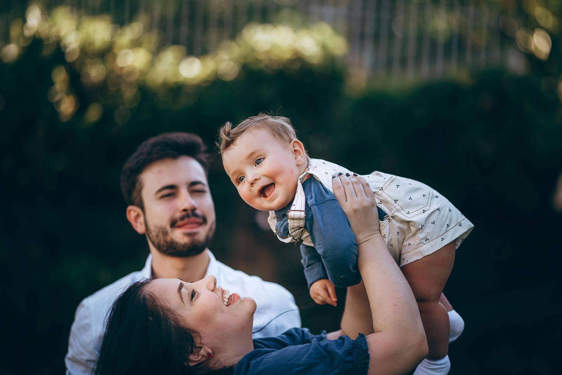 Two adults smiling as they lift a baby outdoors. New parent financial planning, insurance, RESP, cash flow, mortgage.