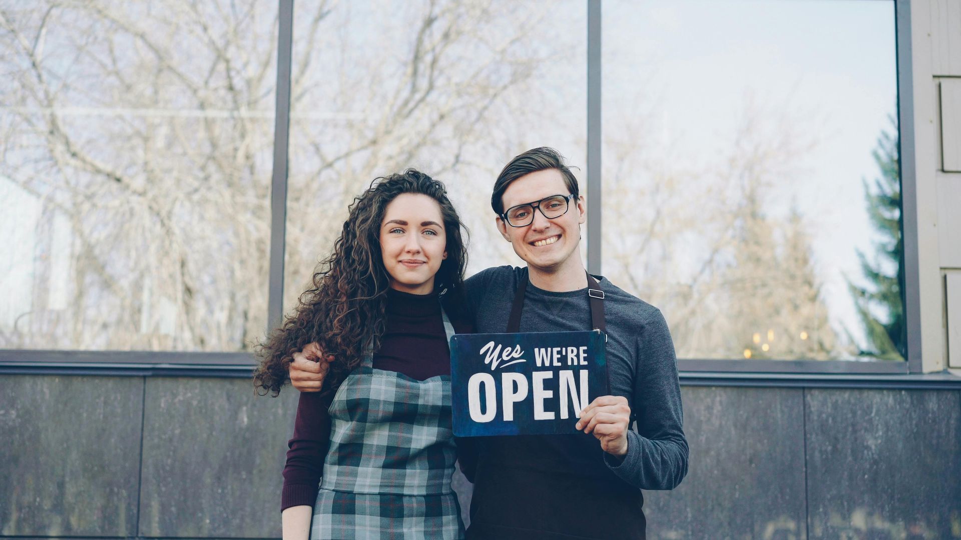 Two people standing outdoors, smiling, holding a sign that says “OPEN”. Business owners, group benefits, corporate insurance, succession planning, retirement strategy