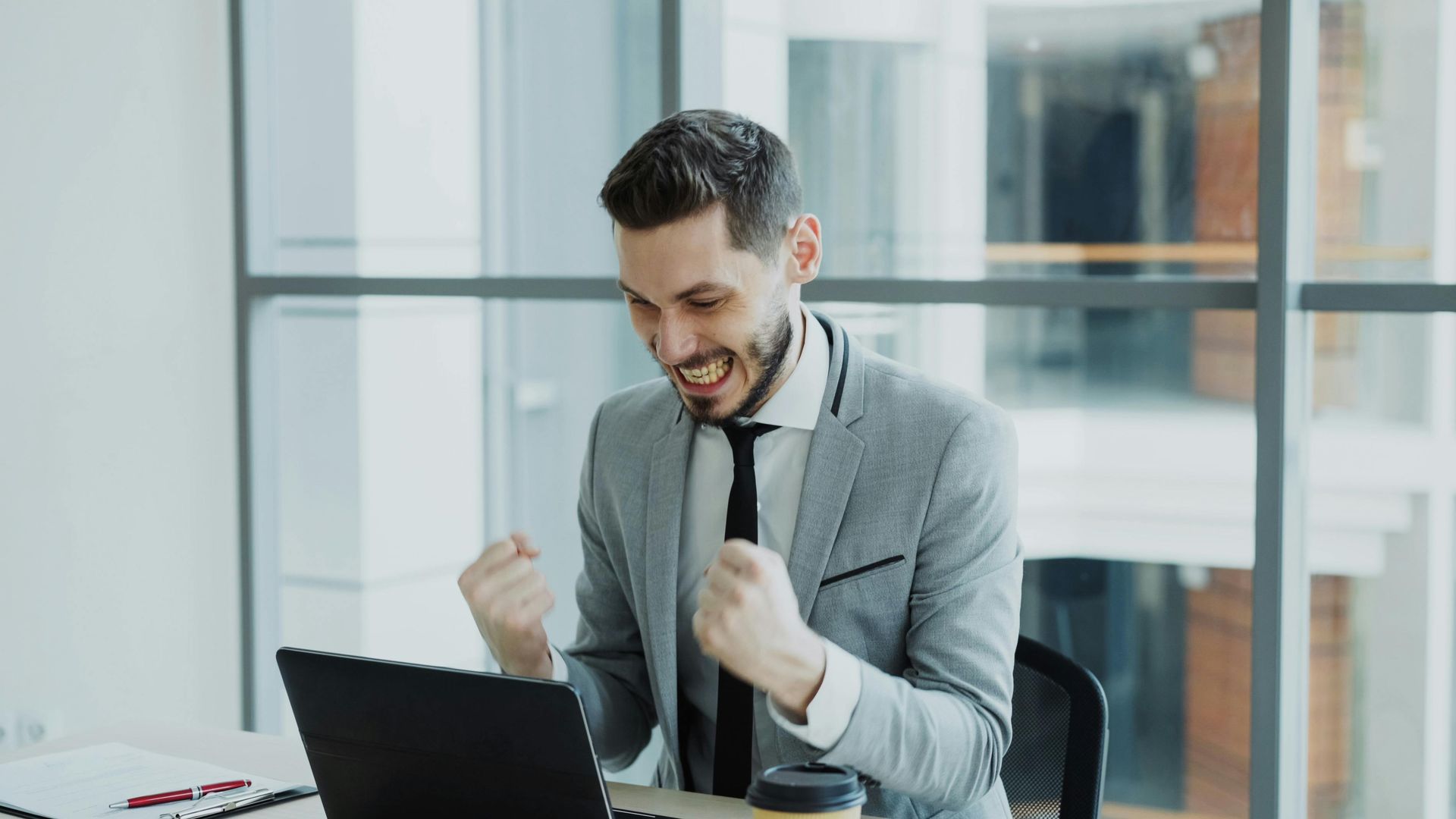 Man in a gray suit celebrating at a laptop in a bright office with clenched fists