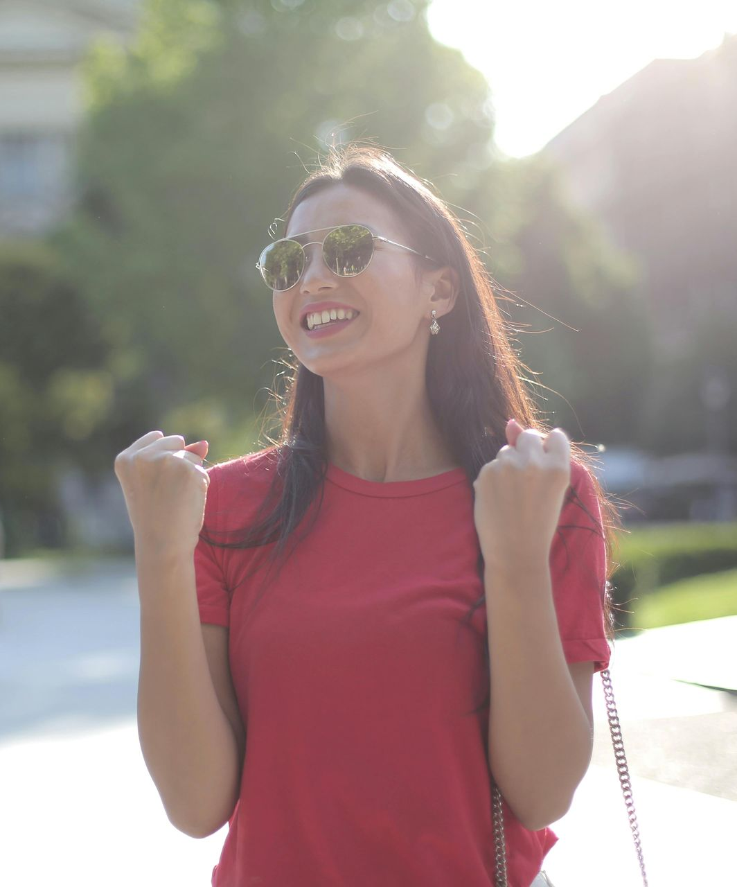 Smiling woman in sunglasses with raised fists, wearing a red shirt outdoors in bright sunlight