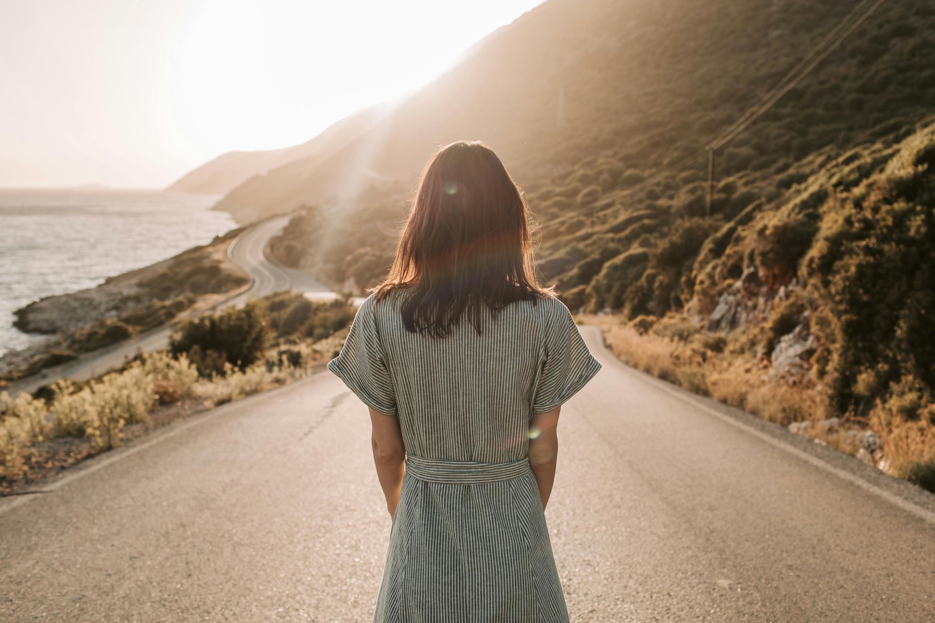 Person stands on a road facing a sunlit coastal hillside and ocean. Unique financial journeys, job loss, divorce, inheritance, new in Canada.