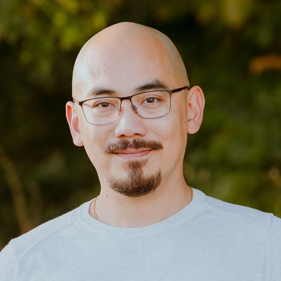 Portrait of a bald man with glasses and a goatee, wearing a light gray shirt, standing outdoors against greenery