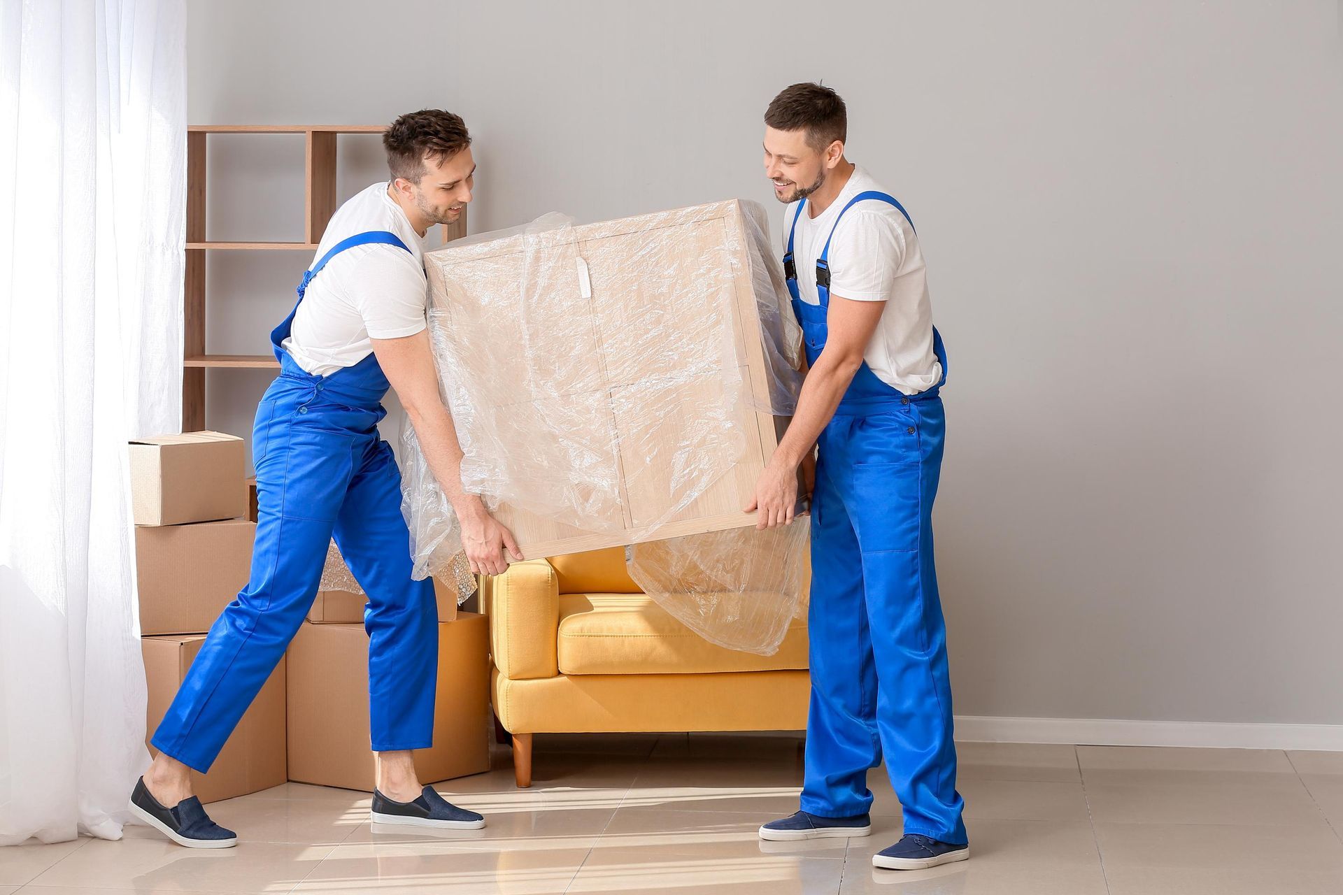 Two movers in blue coveralls carrying a wrapped box in a room with furniture and boxes.