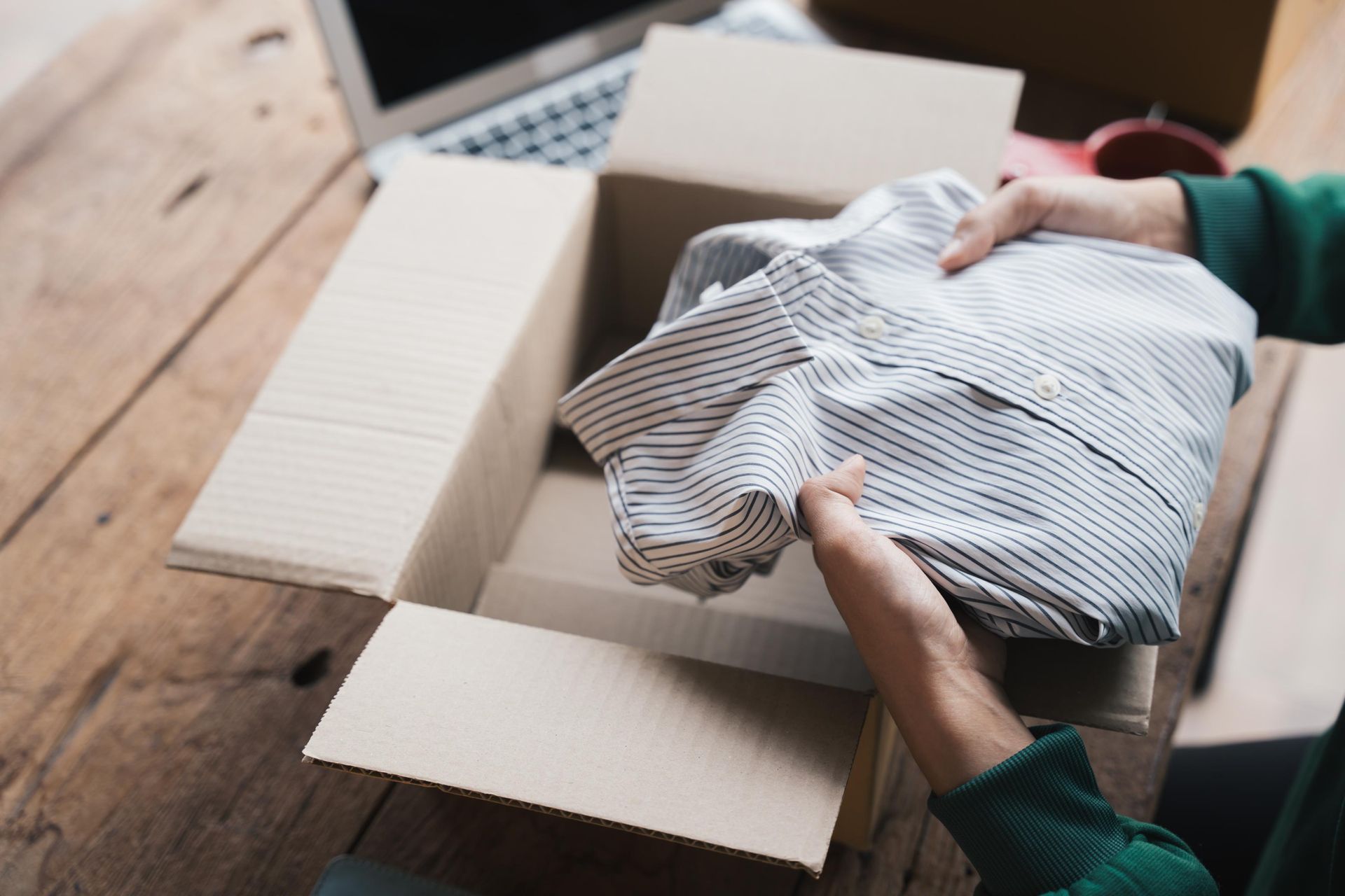 Person unpacking a striped shirt from a cardboard box on a wooden table near a laptop.