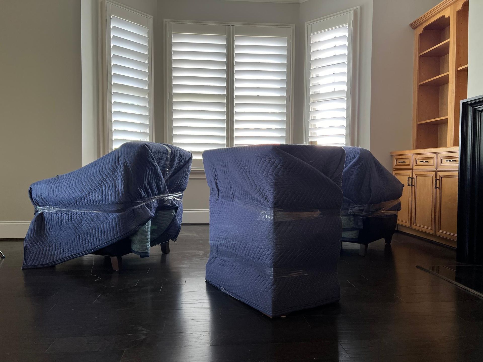 Two upholstered chairs covered in blue moving blankets in a room with hardwood floors and a built-in cabinet.