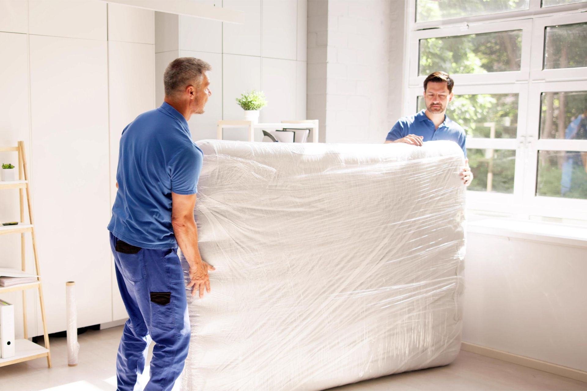 Two men in blue uniforms carry a large, wrapped, bulky item indoors near a window.