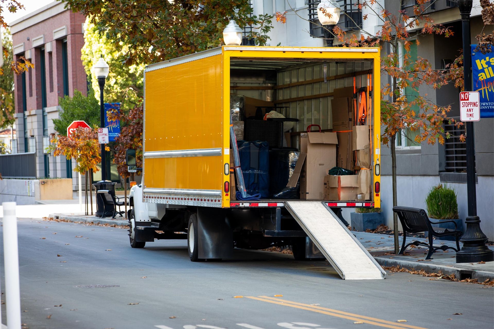 Yellow moving truck parked on a city street, loading ramp down, boxes inside.