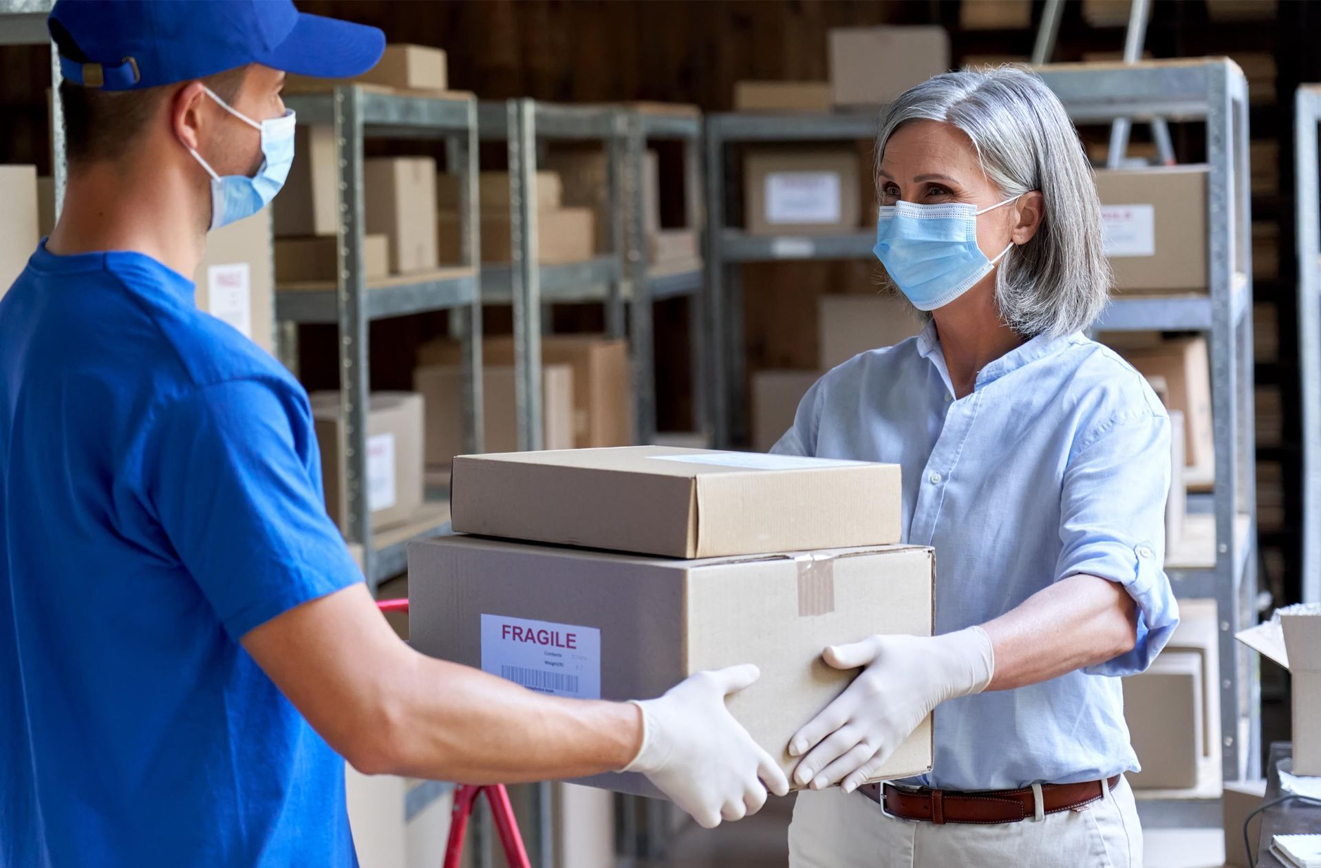 Delivery person in blue mask and uniform handing packages to a woman in a mask, gloves, in a warehouse.