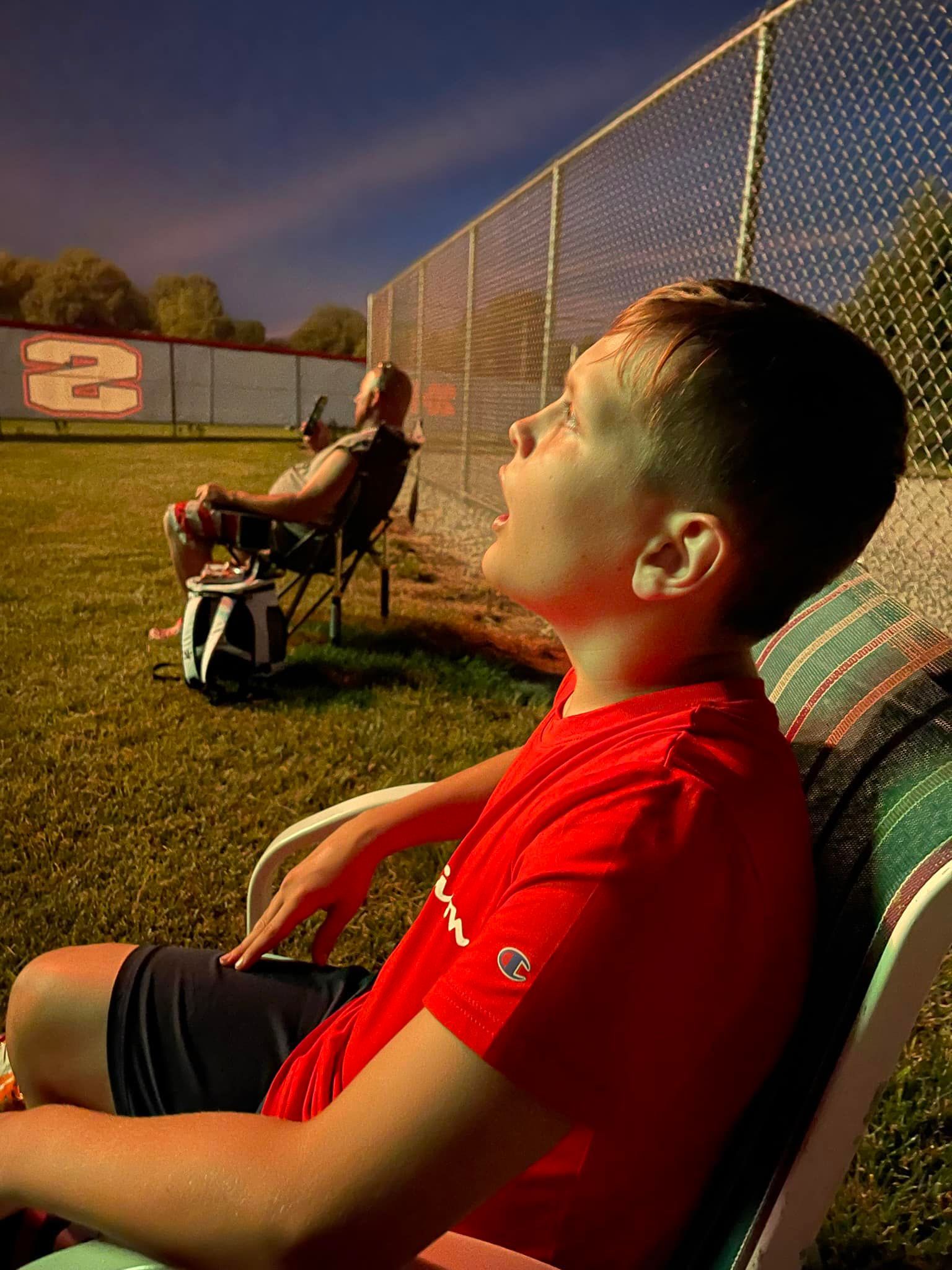 A young boy in a red shirt is sitting in a chair in front of a fence.
