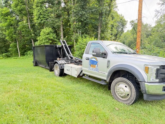 A dump truck is towing a dumpster in a grassy field.