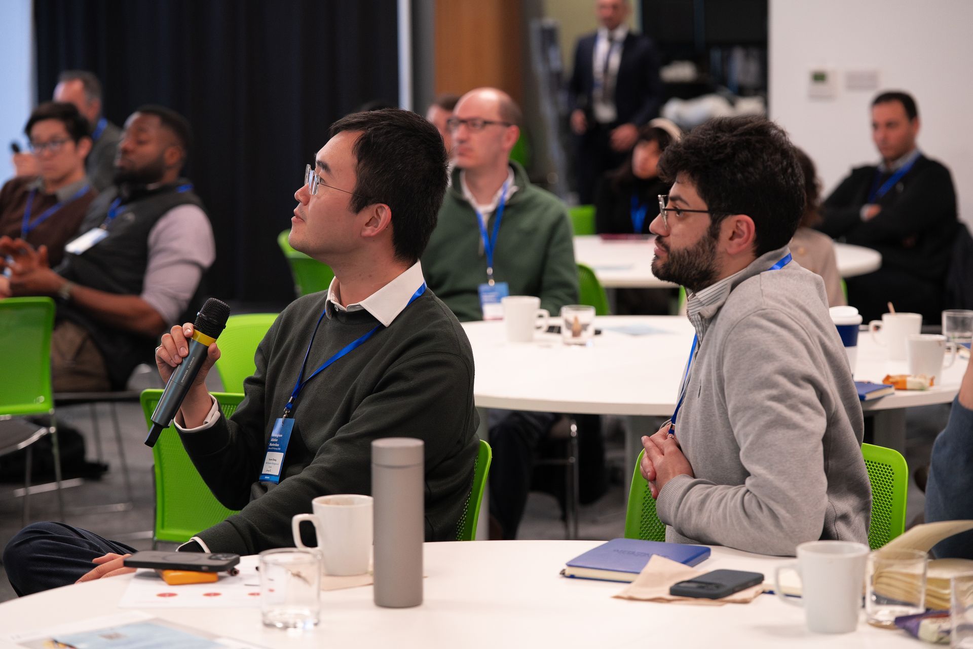 Attendees seated at round tables in a conference room, one person holding a microphone and speaking.