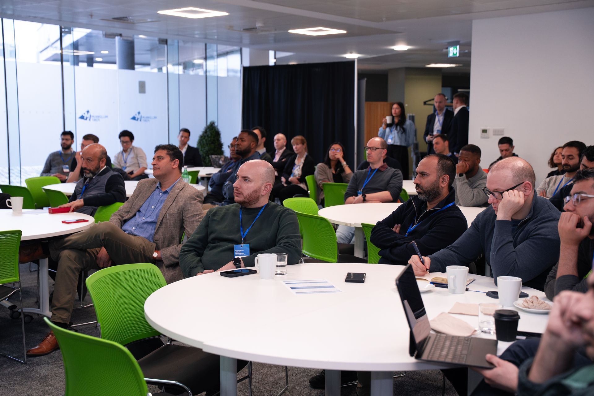 A group of people sit at round tables in a brightly lit office space, attentively listening to a presentation.