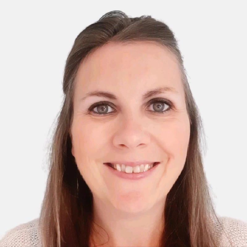A smiling person with long brown hair, wearing a white textured sweater, set against a plain light background.